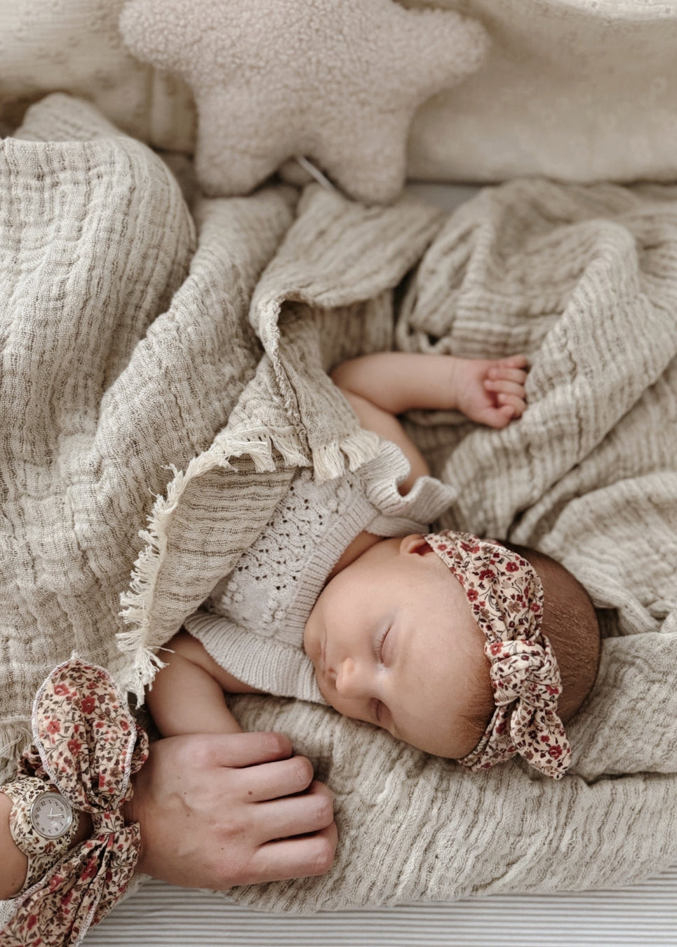 Mãe a dar a mão a um bebé deitado numa cama em tons bejes, ambos a usar acessórios da mrs.ertha no padrão magic garden